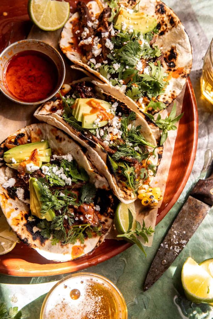 Three grilled tacos filled with meat, avocado slices, fresh herbs, corn, and crumbled cheese are served on a plate with lime wedges—a delicious choice for Mother’s Day brunch ideas—plus red sauce and a glass of beer nearby.