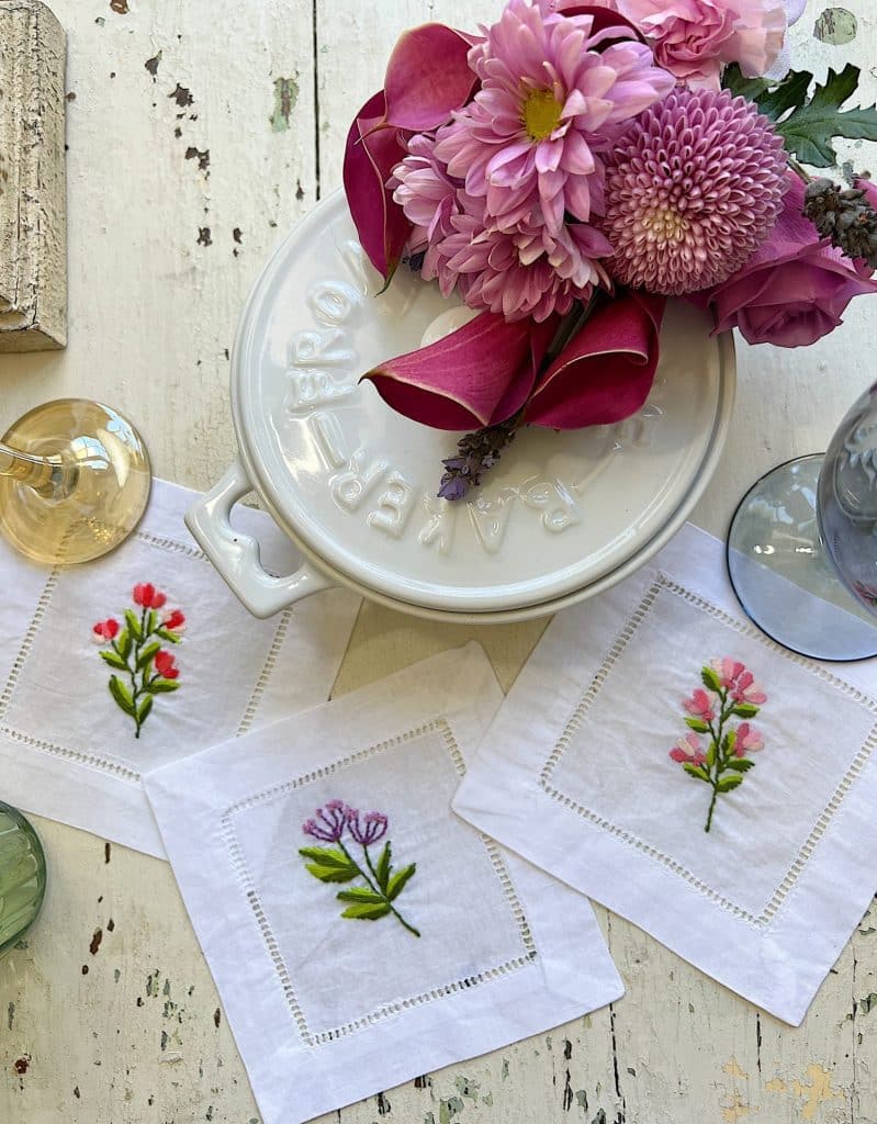 A white table with a round Le Creuset dish, a bouquet of pink flowers, a glass of white wine, a glass of water, and three white napkins embroidered with colorful designs inspired by my favorite flower crafts.