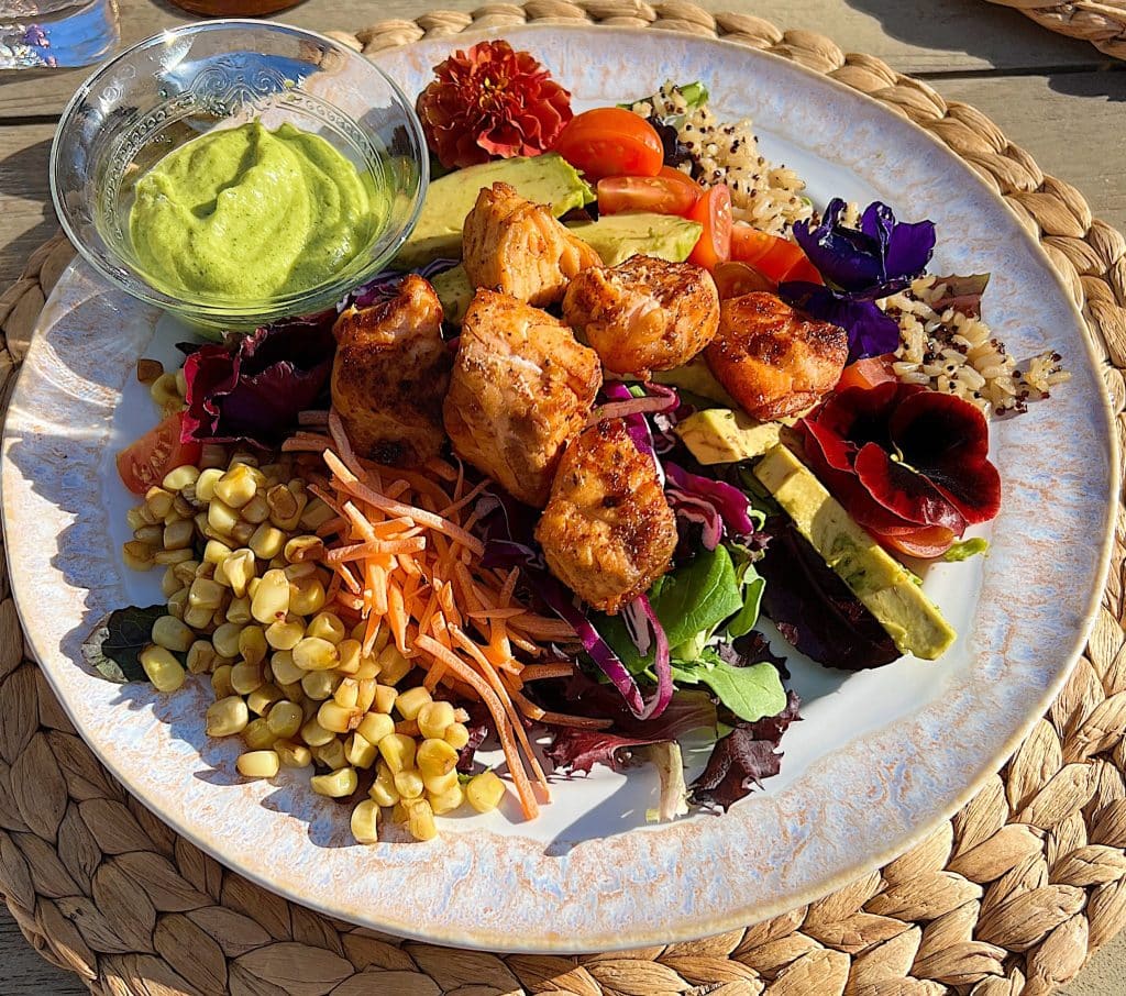 A colorful salad with grilled chicken, avocado, cherry tomatoes, carrots, corn, mixed greens, and edible flowers—served with green sauce on a woven placemat—makes a fresh choice for Mother’s Day brunch ideas.