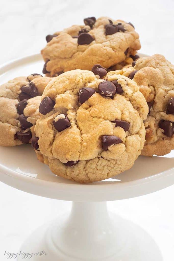 A white cake stand holds a stack of large, thick chocolate chip cookies with golden-brown edges and generous chocolate chips—one of my favorite chocolate recipes—set against a light background.
