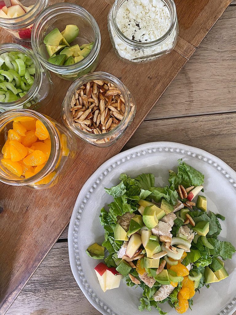 A plate of salad with lettuce, avocado, apple, chicken, pecans, and mandarin oranges—one of my favorite easy dinner ideas—sits next to jars filled with celery, avocado, feta cheese, sunflower seeds, and mandarin oranges on a wooden board.