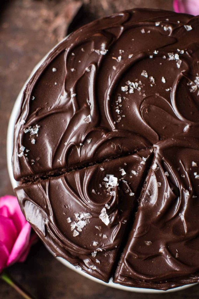 A close-up of a rich, glossy chocolate cake with swirled frosting, topped with flaky sea salt—one of my favorite chocolate recipes. A single slice is partially cut, and pink flowers are visible at the edge of the image.