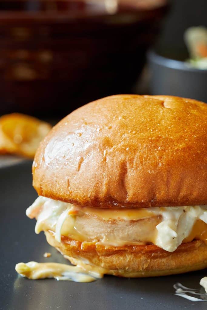A close-up of a chicken sandwich with a golden bun, creamy coleslaw, and chicken fillet, resting on a dark surface with sauce oozing out—one of my favorite easy dinner ideas.