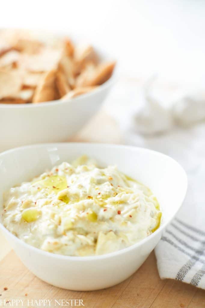 A white bowl filled with creamy artichoke dip, one of my favorite side dishes, is garnished with herbs and sits on a wooden surface. In the background, a bowl of pita chips is slightly out of focus.