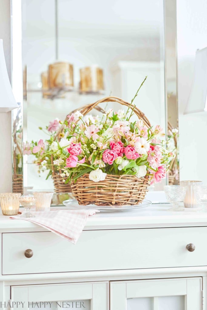 A wicker basket filled with pink and white blooms—my favorite flower inspiration—sits on a white dresser, surrounded by candles and a folded gingham cloth, with a mirror reflecting the charming arrangement in the background.