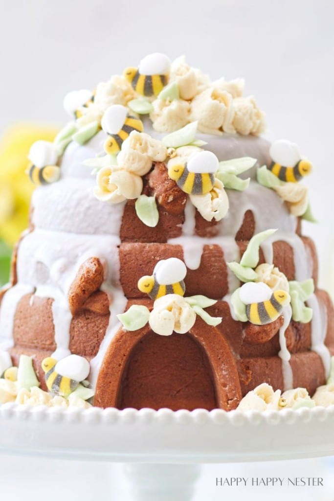 A beehive-shaped cake with white icing drips, decorated with fondant bees, white flowers inspired by my favorite flower inspiration, and green leaves, sits on a white cake stand.