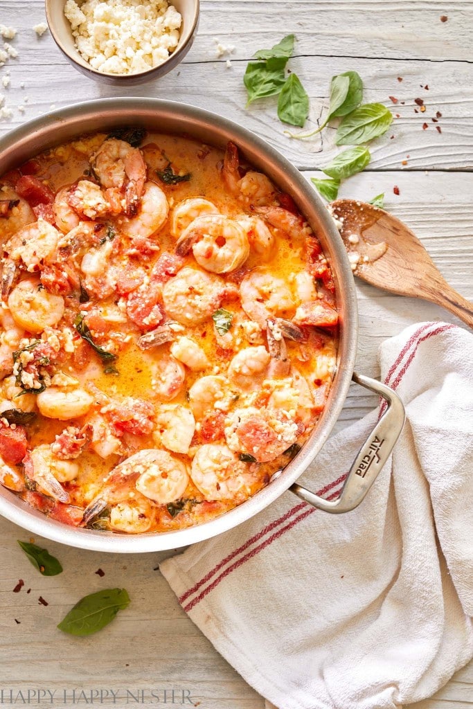 A skillet filled with shrimp in a creamy tomato sauce, garnished with herbs, sits on a white wooden table—one of my favorite easy dinner ideas—next to a white towel, scattered basil leaves, and a small bowl of crumbled cheese.