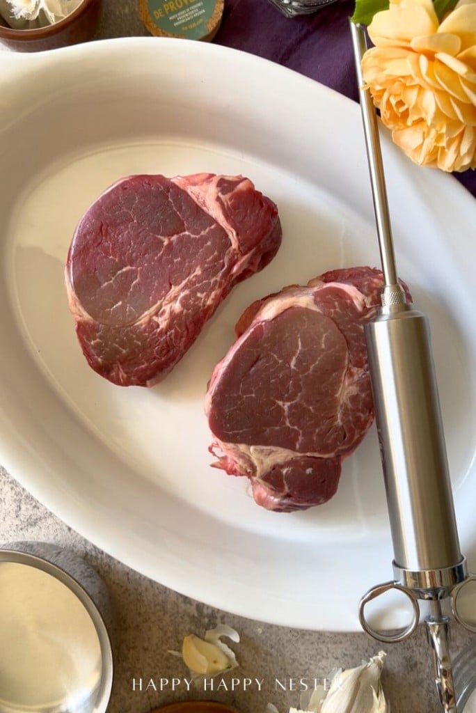Two raw beef steaks are placed on a white oval platter. Next to the platter is a stainless steel meat injector, a bowl, garlic cloves, a flower, and part of a spice container.