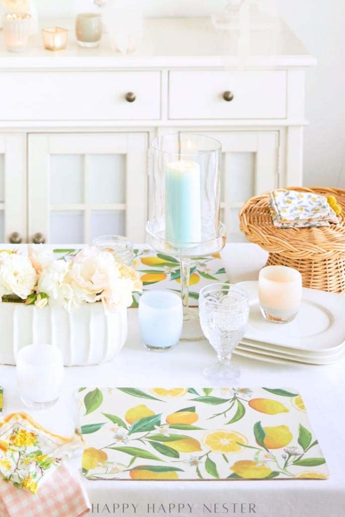 A bright, elegant table setting with easy corkboard placemats, white dishes, crystal glasses, candles, a basket of napkins, and a white floral arrangement on a white table in front of a white cabinet.