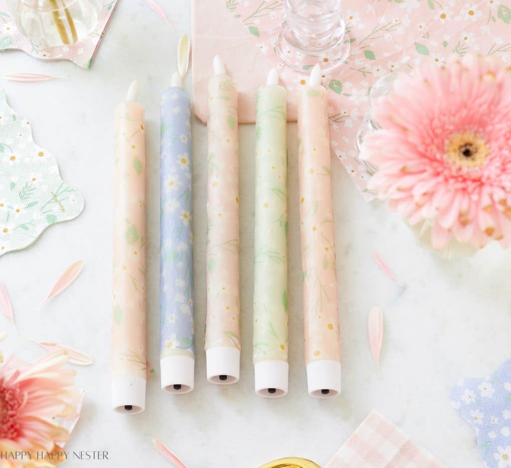 Five pastel-colored flameless candles with floral patterns, inspired by my favorite flower, are arranged side by side on a white surface, surrounded by pink flowers, petals, and decorative napkins in soft spring hues.