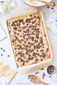 A large tray of freshly baked chocolate chip cookie bars sits on parchment paper, surrounded by kitchen utensils, a bowl with batter, a wooden spoon, a yellow towel, and a small bowl of chocolate chunks on a white surface.