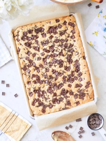 A large tray of freshly baked chocolate chip cookie bars sits on parchment paper, surrounded by kitchen utensils, a bowl with batter, a wooden spoon, a yellow towel, and a small bowl of chocolate chunks on a white surface.