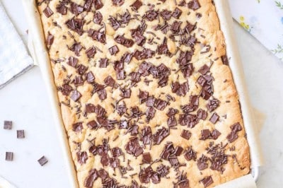 A large tray of freshly baked chocolate chip cookie bars sits on parchment paper, surrounded by kitchen utensils, a bowl with batter, a wooden spoon, a yellow towel, and a small bowl of chocolate chunks on a white surface.
