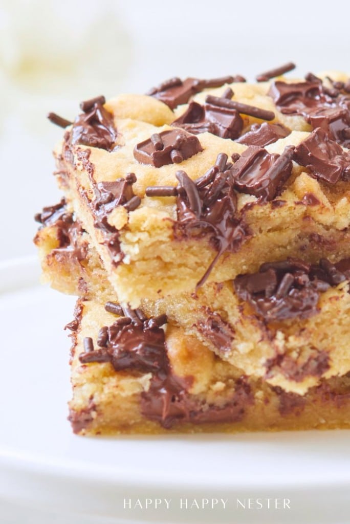Two stacked chocolate chip bars, made using our delicious chocolate chip bars recipe, feature gooey melted chocolate chunks and sprinkles on top. Their golden, chewy texture and rich appearance make them irresistible on a simple white plate.