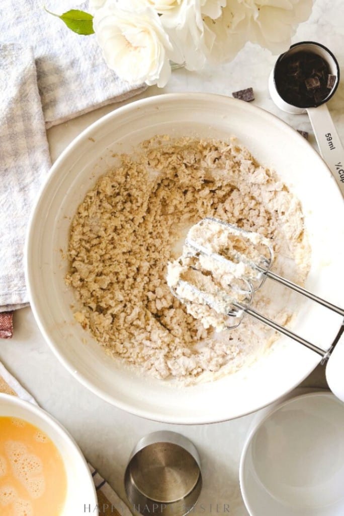 A mixing bowl with cookie dough and an electric hand mixer sits on a countertop, surrounded by ingredients like a bowl of beaten eggs, chocolate chunks, and a metal measuring cup. A towel and white flowers are nearby.
