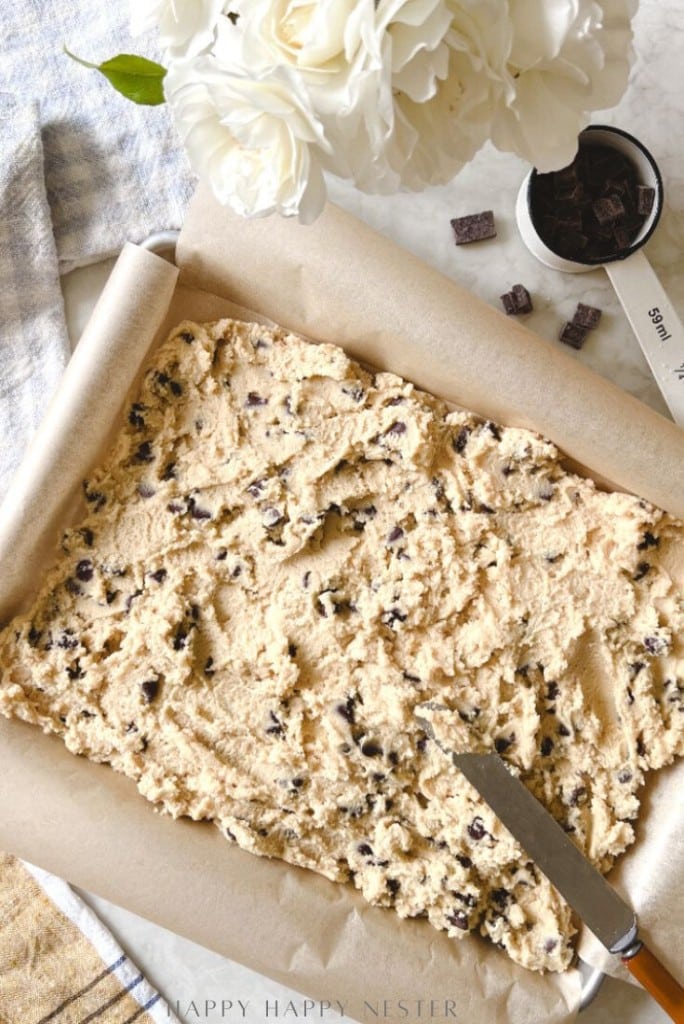 A baking tray lined with parchment paper holds unbaked chocolate chip cookie dough, with a spatula resting on the tray. A white flower in a vase and a measuring cup with chocolate chunks are nearby.