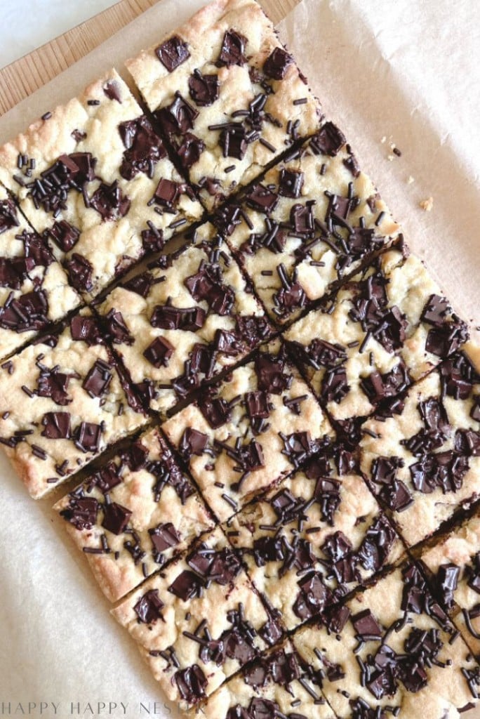 A batch of chocolate chunk cookie bars cut into squares, topped with chunks of chocolate and chocolate sprinkles, resting on parchment paper.