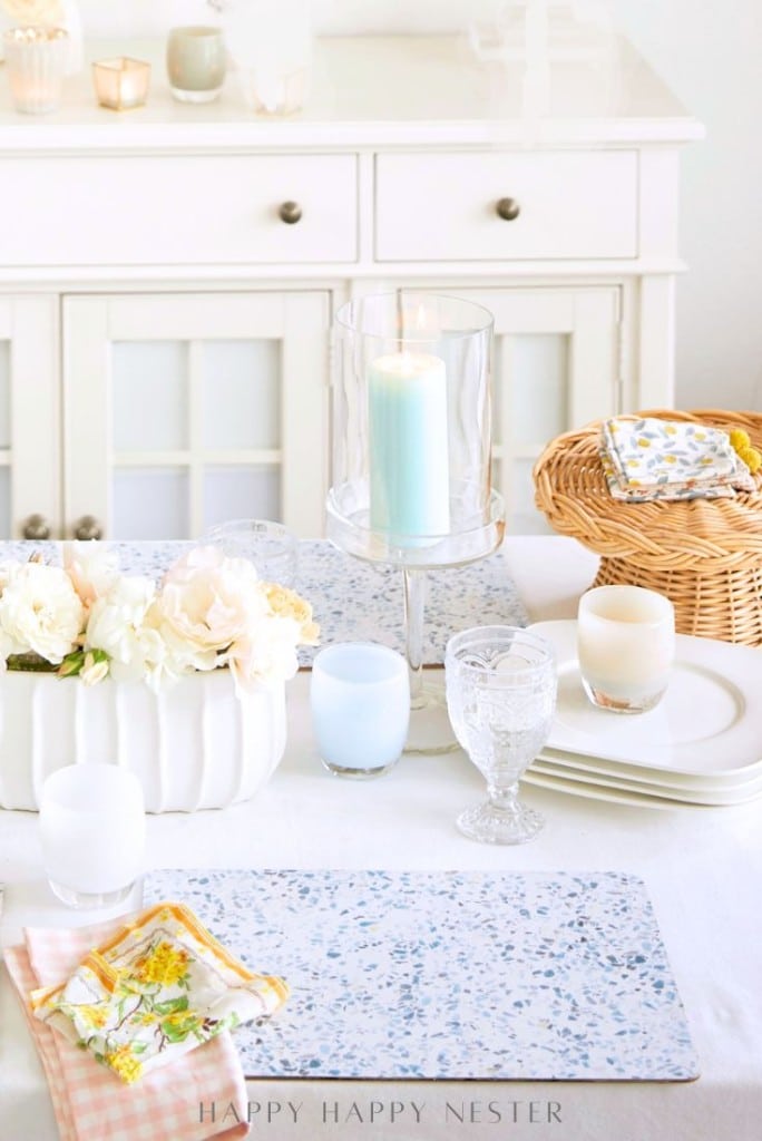 A bright, airy table setting features easy corkboard placemats, floral napkins, white plates, glassware, candles, a basket of napkins, and a centerpiece of white flowers in a ceramic vase on a white tablecloth.