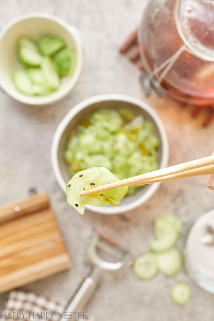 A pair of chopsticks holds a slice of cucumber above a bowl of sliced cucumbers—one of my favorite side dishes—while a cutting board, peeler, and glass teapot sit in the background on a light surface.