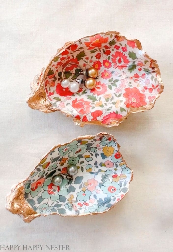 Two oyster shells lined with colorful, my favorite flower-inspired fabric hold pairs of pearl earrings on a light, textured surface. The shells' golden edges complement one red floral pattern and one blue, adding a touch of inspiration to every detail.