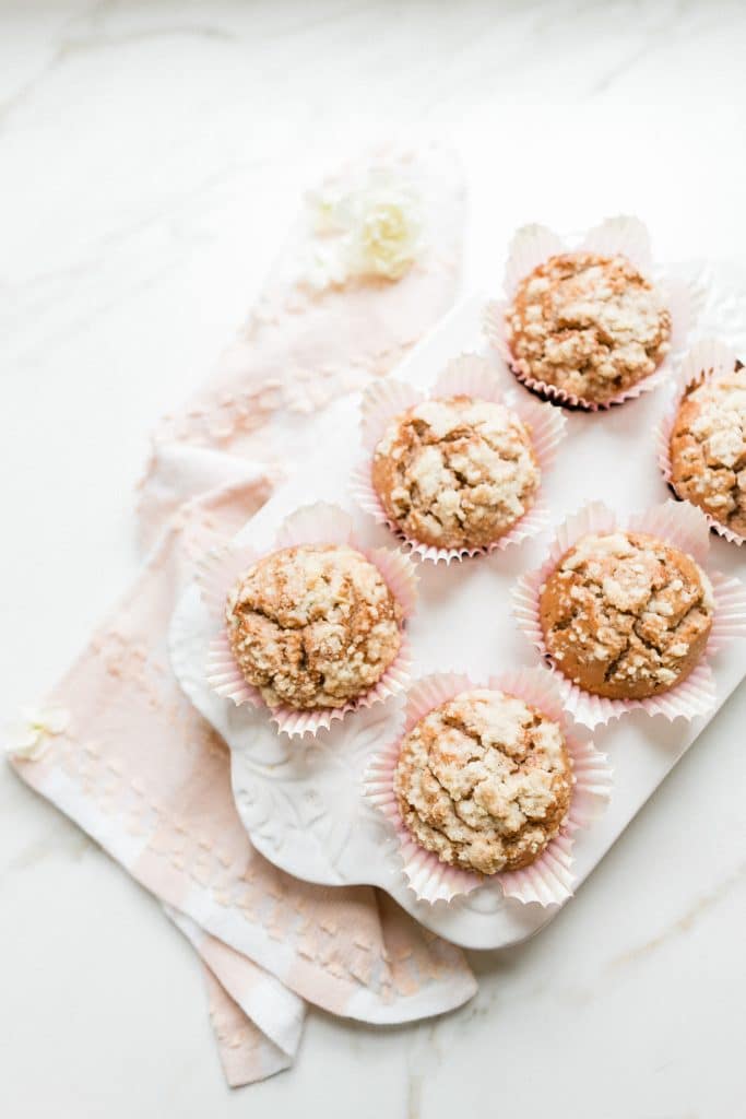 Six muffins with crumbly streusel topping in pink paper liners are arranged on a white tray atop a soft pink cloth—an inviting treat perfect for Mother's Day brunch ideas, set against a white marble surface.