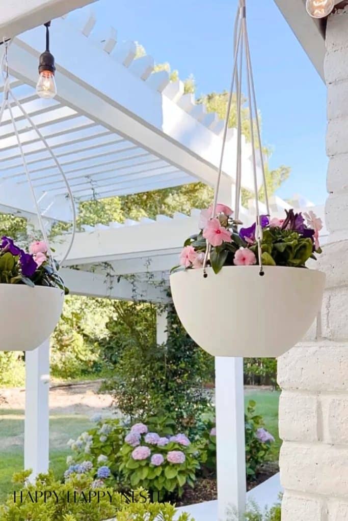 Two white hanging planters filled with pink and purple flowers are suspended on a white pergola, offering my favorite flower inspiration. In the background, blooming hydrangeas and green trees flourish under natural daylight.