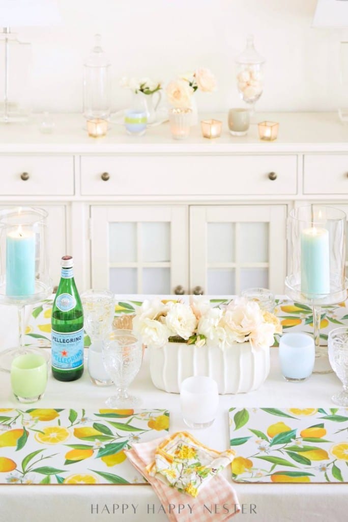 Bright, spring-themed table setting with a floral table runner, white flowers in a ceramic vase, candles in glass holders, Pellegrino water, and elegant glassware on a white dining table and sideboard.