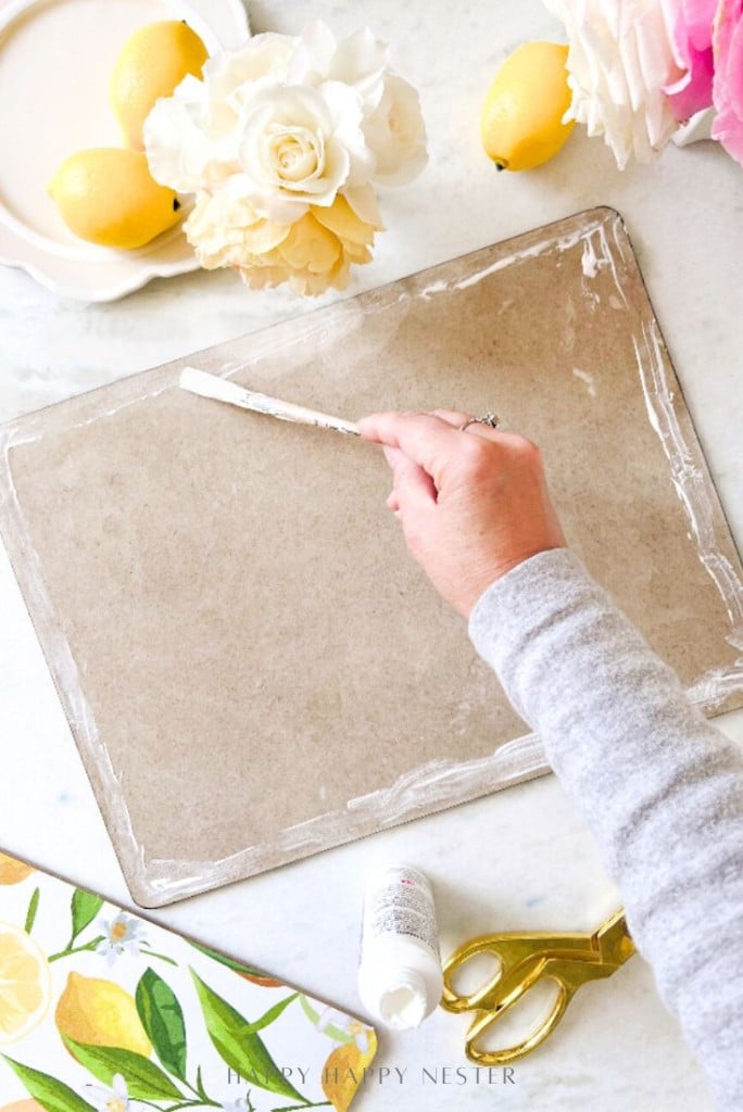 A person spreads white paste onto the edge of a rectangular piece of cardboard. Lemons, flowers, a gold pair of scissors, and a lemon-patterned napkin are on the white table around the workspace.