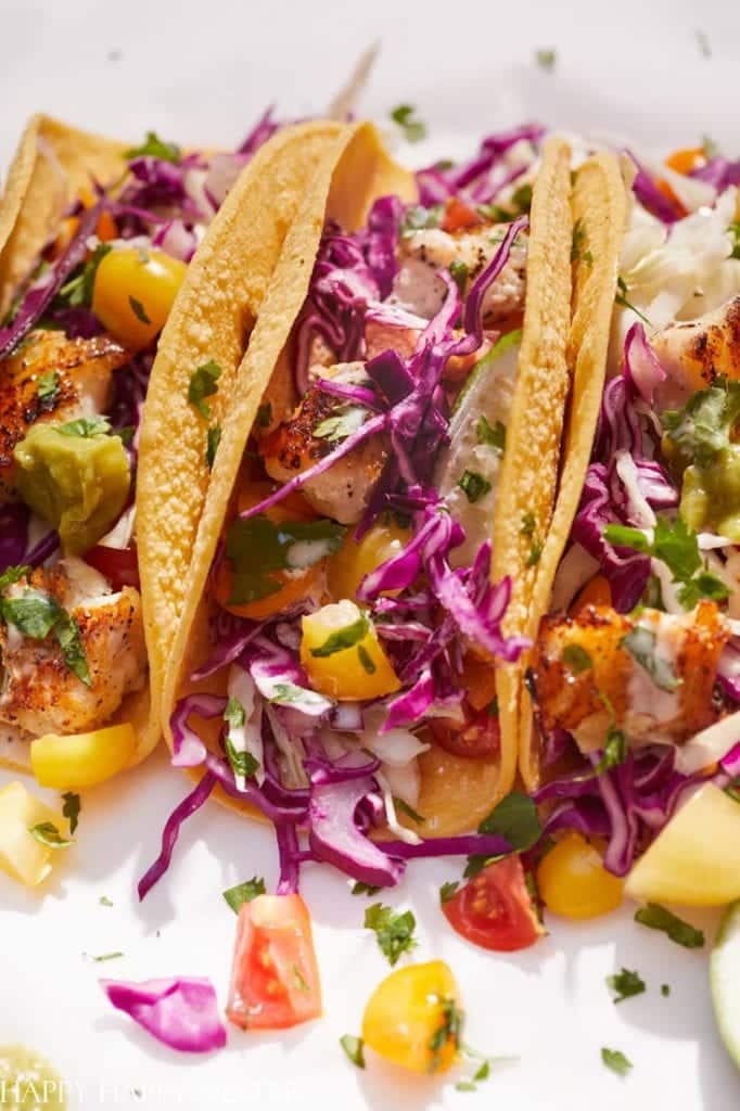 Three fish tacos in corn tortillas topped with shredded purple cabbage, diced tomatoes, avocado, cilantro, and a creamy sauce—one of my favorite easy dinner ideas—served on a white plate.