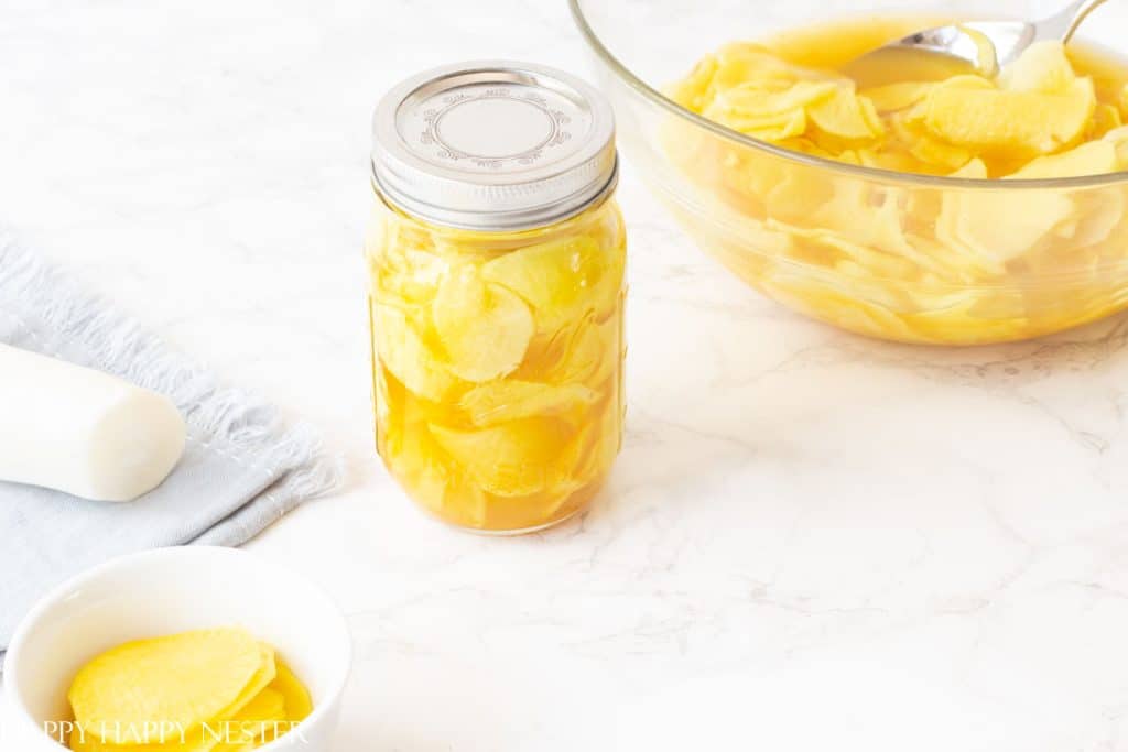 A jar filled with yellow pickled daikon radish slices sits on a marble surface, with a bowl of more pickled radish and a spoon in the background—a classic among my favorite side dishes. A small dish of radish slices is in the foreground.