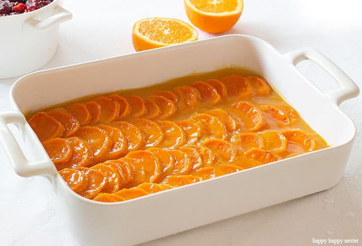 Sliced candied yams arranged in rows in a white baking dish, with an orange half and a bowl of cranberries nearby—one of my favorite side dishes on a bright white surface.