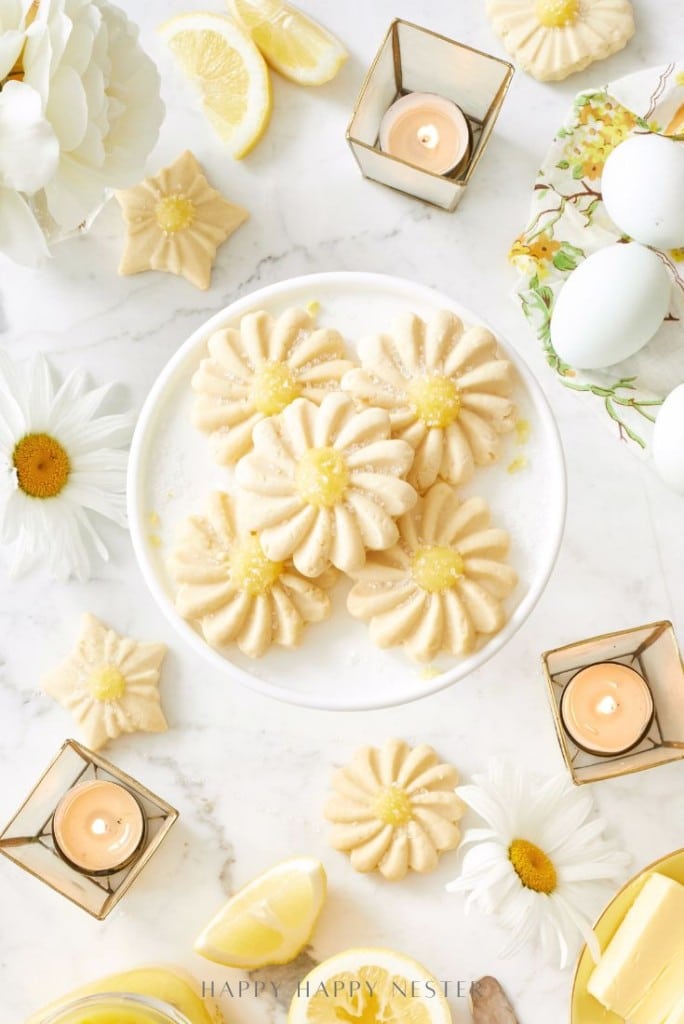A white plate of daisy-shaped lemon cookies sits on a marble surface, surrounded by fresh daisies, lemon slices, eggs, and glowing gold candle holders—an inviting scene for anyone seeking the perfect flower cookies recipe.