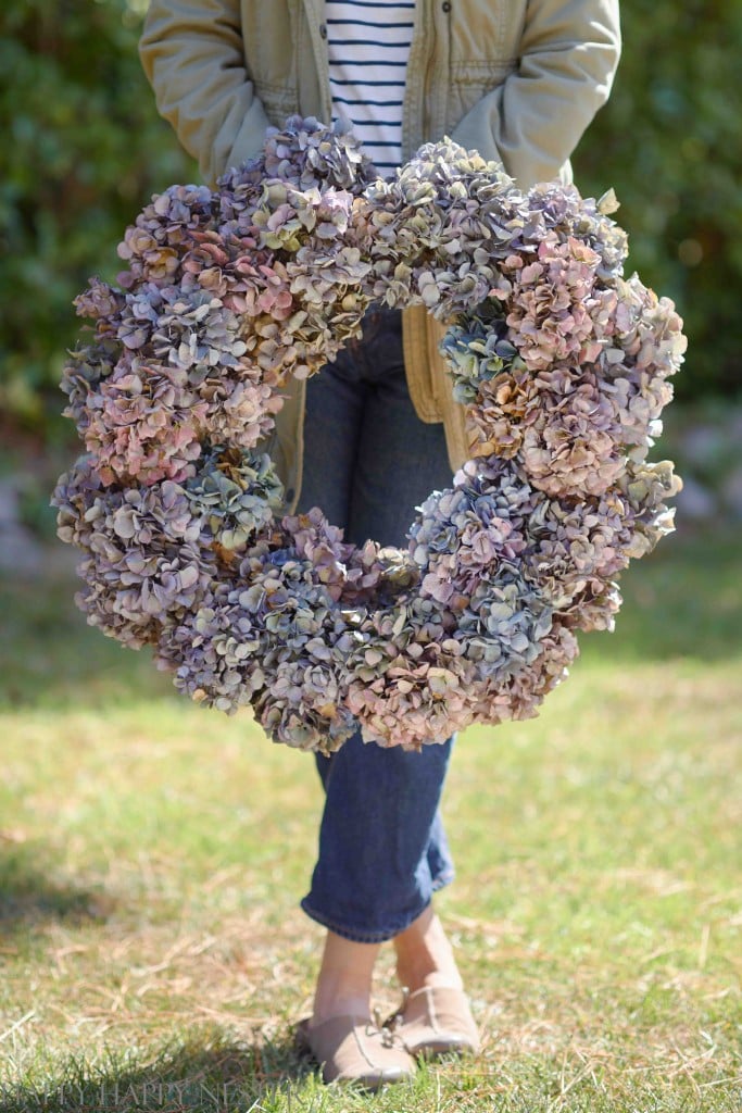 A person in jeans, a striped shirt, and a light jacket stands on grass outdoors, holding a large wreath of dried hydrangea flowers—my favorite flower inspiration for seasonal decor.