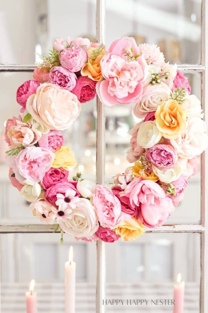 A decorative wreath made of pink, peach, and white artificial flowers—one of my favorite flower crafts—hangs on a window. Three pastel-colored candles are lit in the softly blurred background.