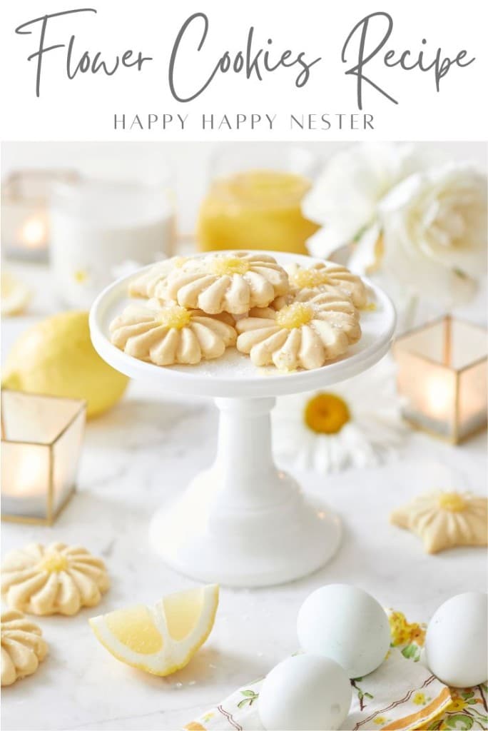 White flower cookies are artfully arranged on a white cake stand, surrounded by candles, lemon slices, flowers, and eggs on marble. The text "Flower Cookies Recipe" completes this elegant scene.