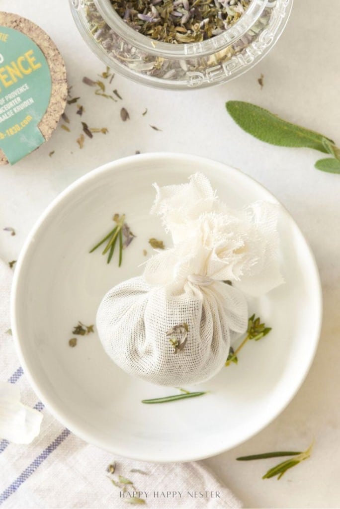 A small mesh pouch filled with dried herbs sits in a white bowl, surrounded by loose herbs and a jar of more dried herbs nearby—perfect ingredients to enhance your instant pot garlic mashed potatoes recipe. A sprig of fresh sage and a cork lid are also visible.