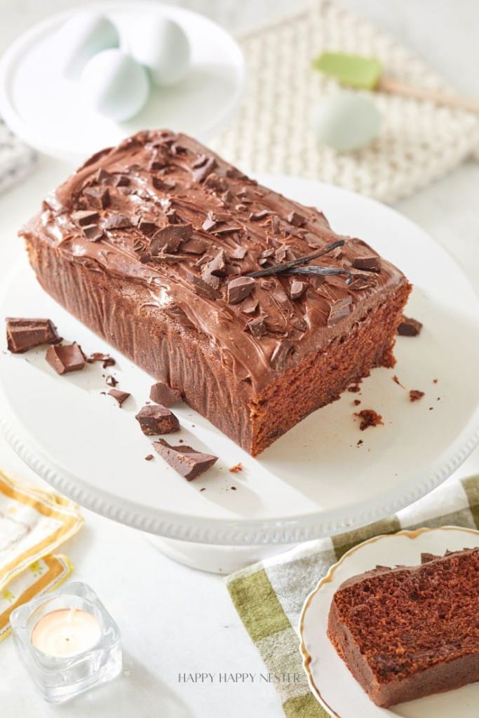 A loaf of chocolate cake—one of my favorite chocolate recipes—topped with frosting and chocolate chunks sits on a white cake stand. Sliced pieces, a candle, and a plate of eggs are nearby on a light-colored table.