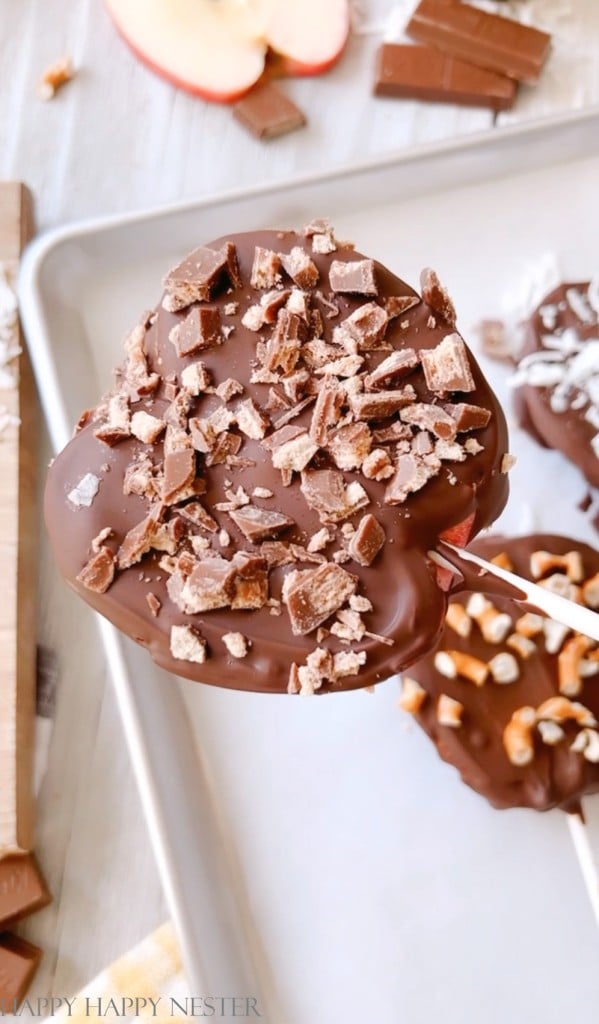 A chocolate-covered apple slice on a stick, topped with chopped toffee pieces, held above a baking tray filled with my favorite chocolate recipes—similar treats and scattered chocolate and apple slices nearby.