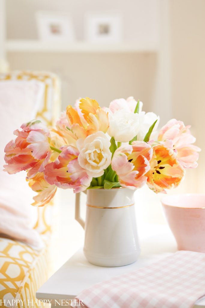 A white ceramic pitcher filled with pink, peach, and white tulips—my favorite flower inspiration—sits on a white table next to a pink bowl and a checked cloth in a bright, cozy room.