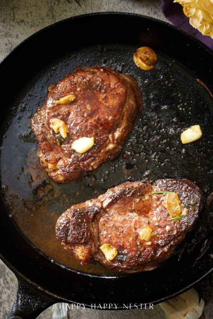 Two cooked steaks in a black cast iron skillet, garnished with sautéed garlic and surrounded by juice and oil—who can resist?