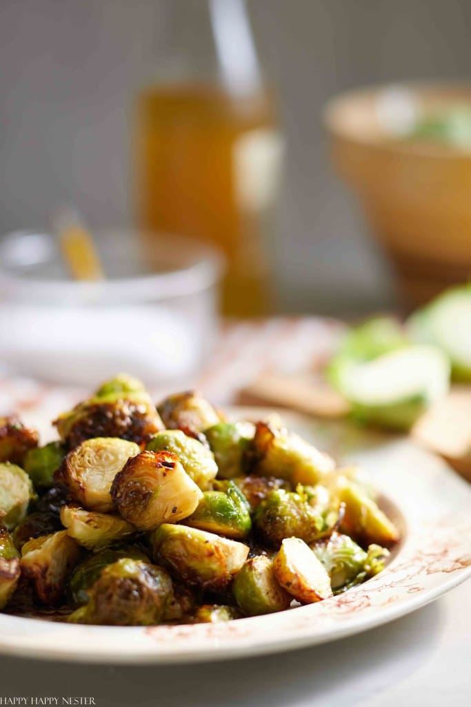 A plate of roasted Brussels sprouts, one of my favorite side dishes, sits on a table, golden brown and lightly garnished. In the blurred background are a bottle, a bowl, and other kitchen items.