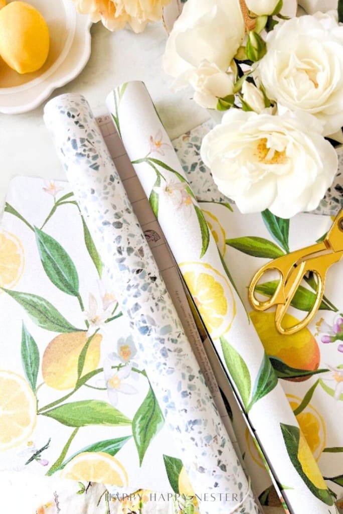 Several rolls of decorative paper with lemon and terrazzo patterns are displayed on a table with white roses, a yellow lemon, and gold scissors nearby—perfect for DIY projects like easy corkboard placemats. The scene is bright and cheerful.