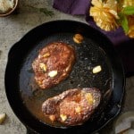 A cast iron skillet with two cooked steaks and garlic cloves sits on a countertop, surrounded by garlic, rosemary, a yellow rose, and a purple cloth—an inviting scene that hints at how to make a tender juicy steak with simple elegance.