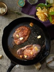 A cast iron skillet with two cooked steaks and garlic cloves sits on a countertop, surrounded by garlic, rosemary, a yellow rose, and a purple cloth—an inviting scene that hints at how to make a tender juicy steak with simple elegance.