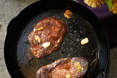 A cast iron skillet with two cooked steaks and garlic cloves sits on a countertop, surrounded by garlic, rosemary, a yellow rose, and a purple cloth—an inviting scene that hints at how to make a tender juicy steak with simple elegance.