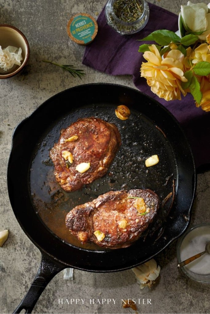 A cast iron skillet with two cooked steaks and garlic cloves sits on a countertop, surrounded by garlic, rosemary, a yellow rose, and a purple cloth—an inviting scene that hints at how to make a tender juicy steak with simple elegance.