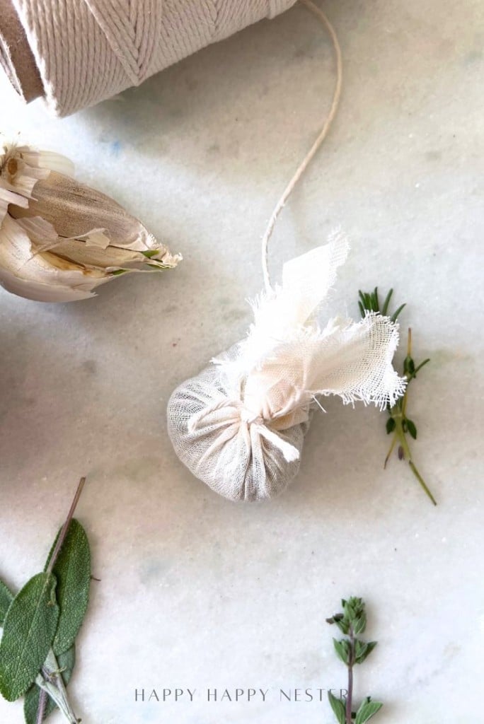 A small herb sachet wrapped in white fabric and tied with string sits on a marble surface, surrounded by fresh herbs and a spool of twine.