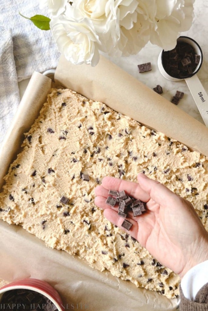 A hand holds chunks of chocolate above a pan filled with unbaked cookie dough—perfect for a chocolate chip bars recipe. Nearby, a cup of chocolate chunks and white flowers rest on the marble countertop.
