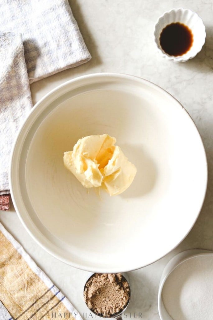 A large white bowl with a scoop of butter sits on a countertop—ready for your chocolate chip bars recipe. Nearby are a bowl of vanilla extract, ramekin of brown sugar, granulated sugar, and folded kitchen towels.