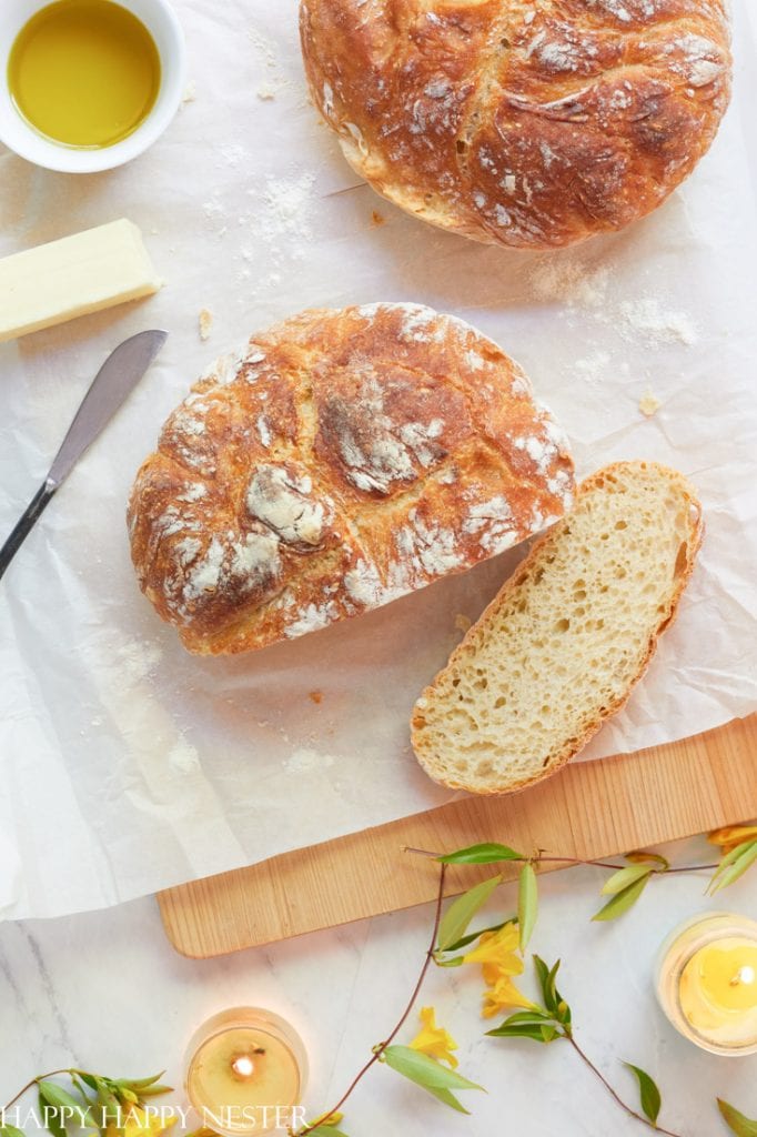 A round loaf of artisan bread, one slice cut, sits on parchment paper next to a butter knife, a stick of butter, a small bowl of olive oil—my favorite side dishes—candles, and yellow flowers on a light table.